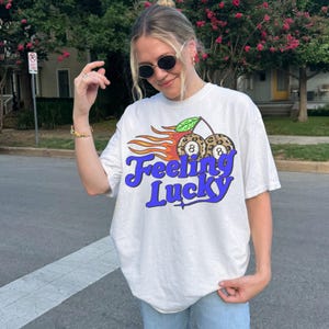K&ouml;nnte beinhalten: Wei&szlig;es T-Shirt mit einem Grafikdesign mit zwei Kirschen mit Leopardenmuster, jeweils mit einer Achtkugel und Flammen. Der Text "Feeling Lucky" ist in blauer Schrift.