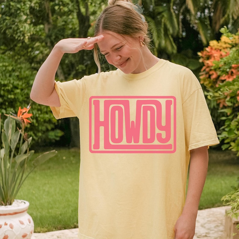 May include: A pale yellow t-shirt featuring the word "HOWDY" in a pink, block-letter design. The shirt is worn by a person outdoors, with a backdrop of green foliage and a potted plant with orange flowers.