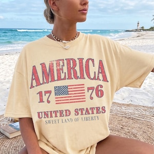 May include: A pale yellow t-shirt featuring the word "AMERICA" in red, the number "1776", and a small American flag graphic. The text "UNITED STATES SWEET LAND OF LIBERTY" is printed below. The shirt is worn on a beach.