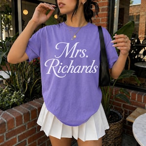 Może przedstawiać: Fioletowy t-shirt z kr&oacute;tkim rękawem z białym napisem Mrs. Richards wydrukowanym kursywą na klatce piersiowej. Koszulka jest zestawiona z białą plisowaną sp&oacute;dnicą i czarną torbą na ramię, z założonym na szyi złotym naszyjnikiem.