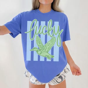 May include: A blue t-shirt with the word "Lucky" in green script above a green duck graphic. The duck is centered on a light blue and white striped background. The shirt has short sleeves.