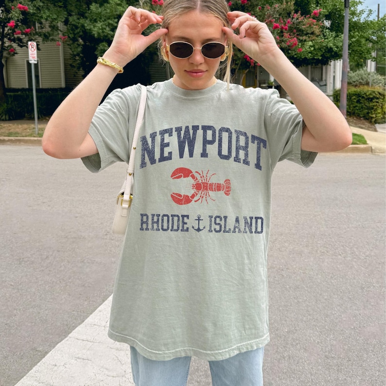 May include: Sage green t-shirt with the words "NEWPORT" and "RHODE ISLAND" printed in navy blue. A red lobster graphic is centered between the words. The shirt is a casual, oversized fit.