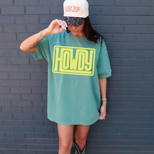 May include: A teal t-shirt with the word "HOWDY" in a yellow, rectangular design. The person is wearing a cream baseball cap with the words "I LOVE YOU" and a leopard print brim. The person is also wearing bracelets and cowboy boots.