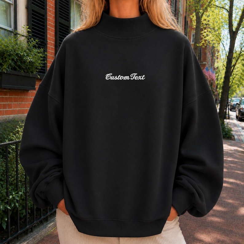 May include: A black long-sleeve sweatshirt with a mock neck collar. The center of the chest features the white embroidered text "CustomText" in a cursive script font. The garment has a relaxed fit with dropped shoulders and ribbed cuffs.
