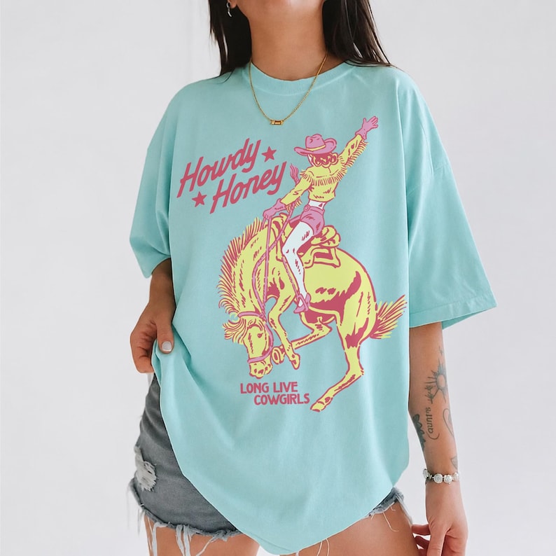 May include: A turquoise t-shirt with a graphic of a cowgirl riding a bucking bronco. The text "Howdy Honey" and "Long Live Cowgirls" are printed on the shirt in pink and yellow. The shirt has short sleeves and a relaxed fit.