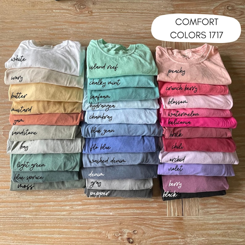 May include: A selection of Comfort Colors 1717 t-shirts in a range of hues, such as white, peachy, and blue jean. Each stack is labelled with the colour name in handwritten script. The image is taken from a top-down perspective.
