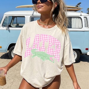 May include: Beige t-shirt with the word "LATER" in pink gingham print and a green alligator graphic. The shirt is a casual, oversized style, perfect for a relaxed look. A surfboard is on top of a blue and white van in the background.