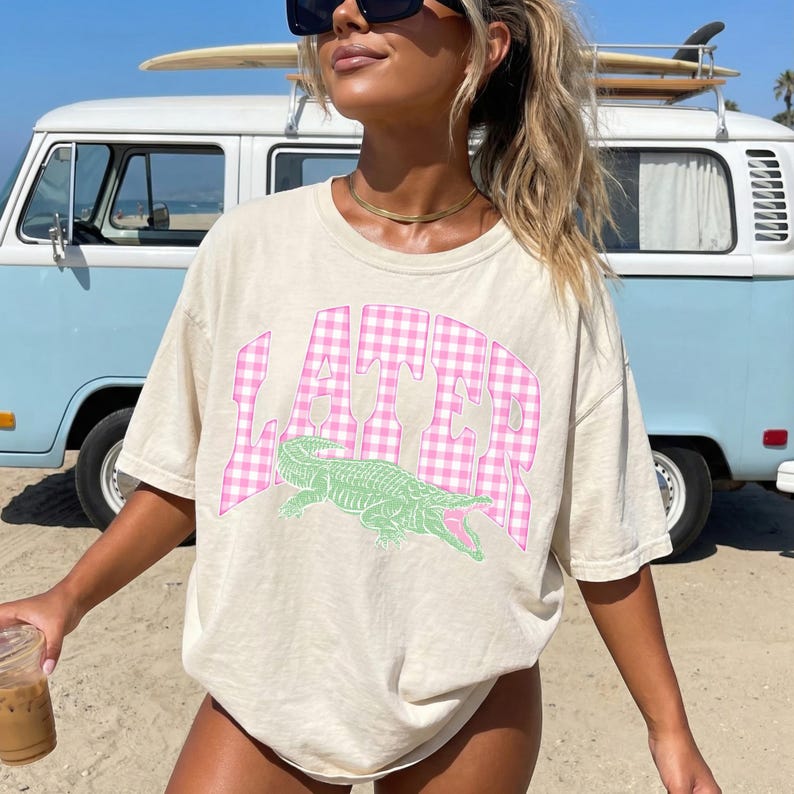 May include: Beige t-shirt with the word "LATER" in pink gingham print and a green alligator graphic. The shirt is a casual, oversized style, perfect for a relaxed look. A surfboard is on top of a blue and white van in the background.