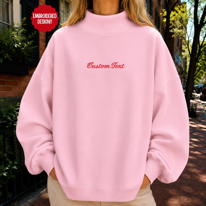 May include: A light pink mock neck sweatshirt featuring long sleeves and a relaxed fit. The center of the chest displays the red embroidered text Custom Text. A red circular badge in the upper left corner contains the text EMBROIDERED DESIGN.