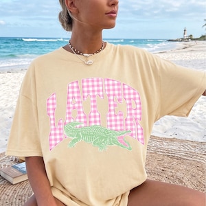 May include: A light yellow t-shirt with the word "LATER" in pink gingham lettering and a green alligator graphic. The shirt is oversized and has short sleeves. The background is a beach scene.