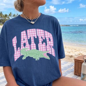 May include: Navy blue t-shirt with the word "LATER" in pink gingham lettering and a green alligator graphic. The shirt is a relaxed fit and is being worn on a beach.