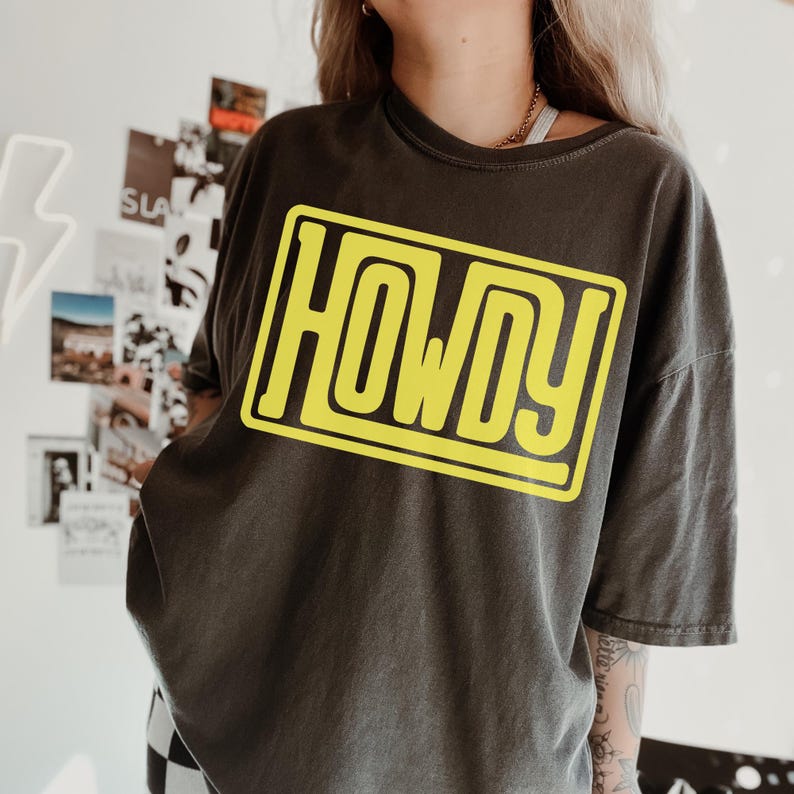 May include: Dark grey t-shirt featuring the word "HOWDY" in bold yellow lettering within a yellow rectangle. The shirt has short sleeves and a relaxed fit. The fabric appears soft and comfortable.