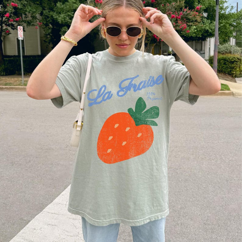 May include: Sage green t-shirt featuring a large orange strawberry graphic and the text "La Fraise" in blue script. The shirt also includes the text "100% Organic". The t-shirt is a relaxed fit.