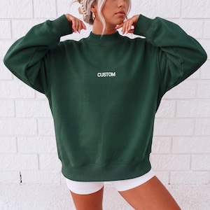 May include: A dark forest green long-sleeve sweatshirt featuring a mock neck collar and ribbed cuffs. The word CUSTOM is printed in white capital letters across the center of the chest. The garment has a loose, relaxed fit with dropped shoulders.