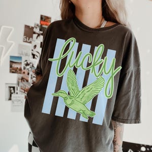 May include: Dark gray t-shirt with a green duck graphic and the word "Lucky" in green script. The design includes light blue vertical stripes. The shirt is a casual, oversized style.