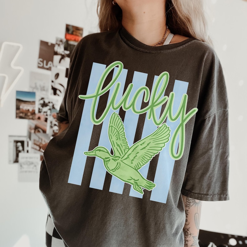 May include: Dark gray t-shirt with a green duck graphic and the word "Lucky" in green script. The design includes light blue vertical stripes. The shirt is a casual, oversized style.