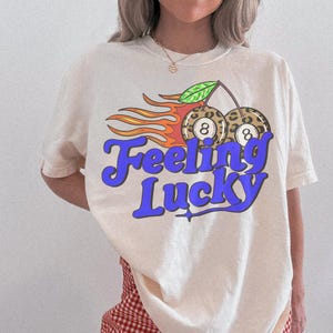 K&ouml;nnte beinhalten: Cremefarbenes T-Shirt mit Retro-Design. Die Grafik zeigt zwei 8-Ball-Kirschen im Leopardenmuster mit Flammen und dem Text "Feeling Lucky" in Blau. Ein lustiges und stilvolles T-Shirt f&uuml;r den l&auml;ssigen Gebrauch.