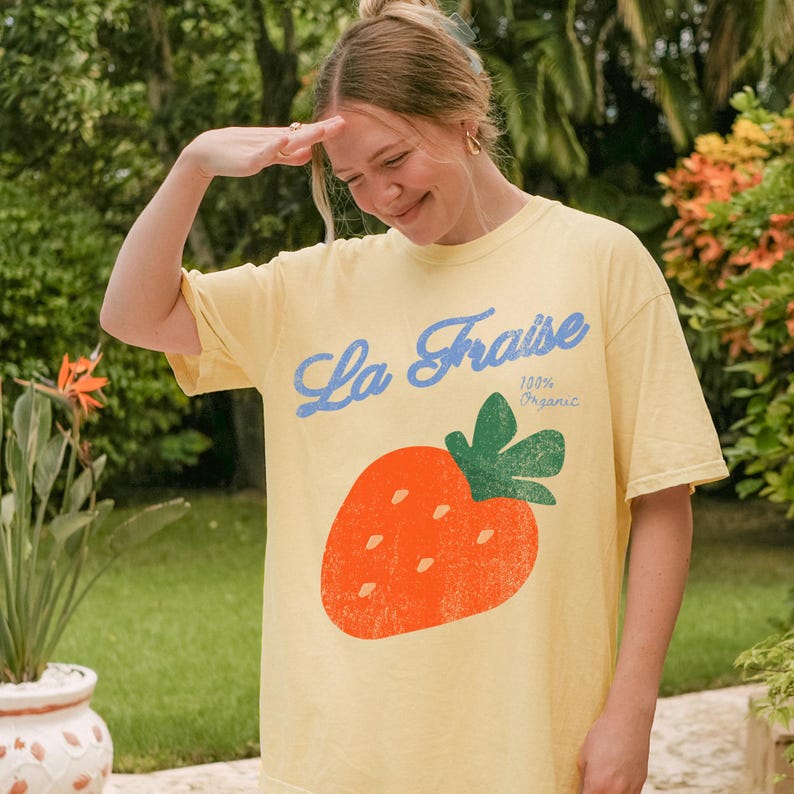 May include: A pale yellow t-shirt featuring a large orange strawberry design and the words "La Fraise 100% Organic" in blue and green. The shirt is worn by a person outdoors.