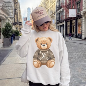 May include: White sweatshirt featuring a watercolor illustration of a teddy bear wearing a brown sweater with a logo design. The bear has a friendly expression. The person is wearing a tan and gray baseball cap with text.