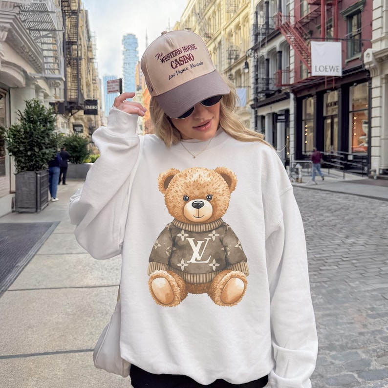 May include: White sweatshirt featuring a watercolor illustration of a teddy bear wearing a brown sweater with a logo design. The bear has a friendly expression. The person is wearing a tan and gray baseball cap with text.