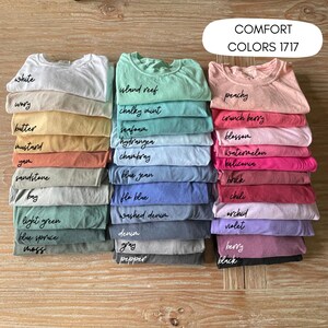 K&ouml;nnte beinhalten: Gestapelte Comfort Colors 1717 T-Shirts in verschiedenen Farben, darunter Wei&szlig;, Pfirsich und Blue Jean. Jeder Stapel ist mit dem Farbnamen in handschriftlicher Schrift beschriftet. Die Aufnahme erfolgt aus der Vogelperspektive.