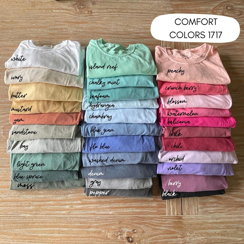 K&ouml;nnte beinhalten: Gestapelte Comfort Colors 1717 T-Shirts in verschiedenen Farben, darunter Wei&szlig;, Pfirsich und Blue Jean. Jeder Stapel ist mit dem Farbnamen in handschriftlicher Schrift beschriftet. Die Aufnahme erfolgt aus der Vogelperspektive.