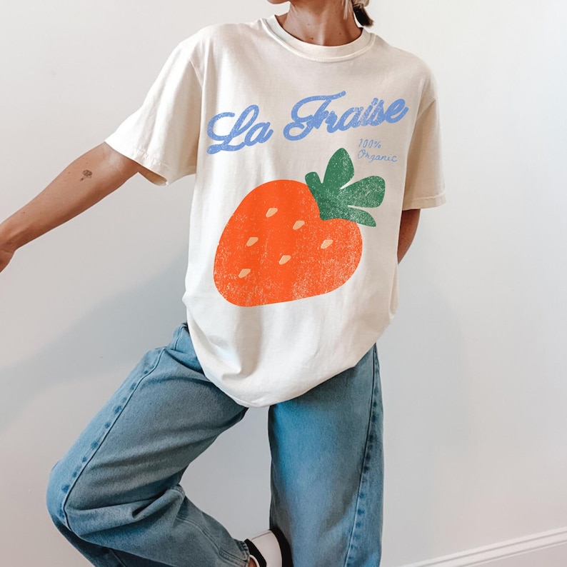 May include: Cream-coloured t-shirt with a large orange strawberry graphic and green leaves. The shirt has the text "La Fraise" in blue script above the strawberry and "100% Organic" below it. The t-shirt is made of a soft material.