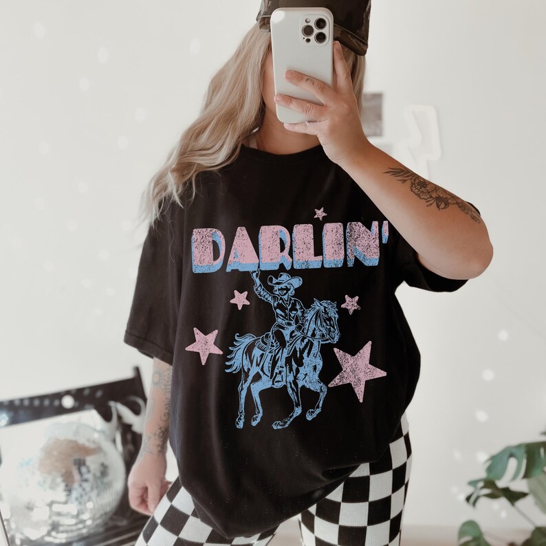Puede incluir: Camiseta negra con la palabra "DARLIN'" en rosa y azul, y un gr&aacute;fico de un vaquero a caballo, rodeado de estrellas rosas. La camiseta tiene un aspecto vintage y desgastado.