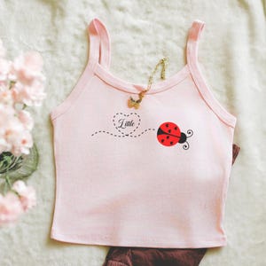 May include: A light pink tank top with a ladybug graphic and the word "Little" inside a heart. A gold butterfly necklace is draped over the top. Brown pants are visible at the bottom of the image. The top is made of a soft material.