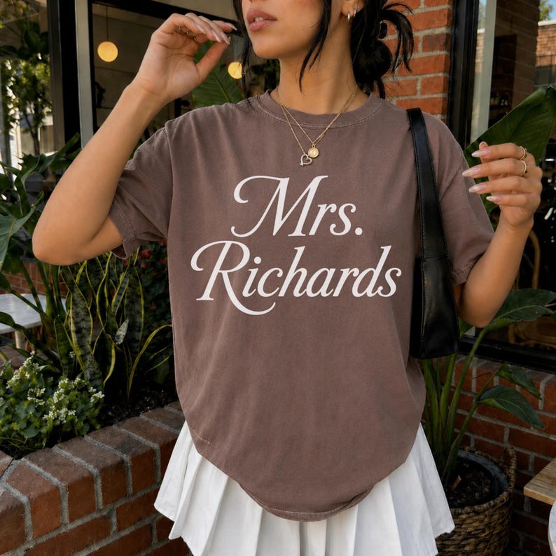 Może przedstawiać: Brązowy t-shirt z kr&oacute;tkim rękawem z białym napisem Mrs. Richards wydrukowanym kursywą na klatce piersiowej. Odzież ma okrągły dekolt i luźny kr&oacute;j, zestawiona z białą plisowaną sp&oacute;dnicą i czarną torbą na ramię.