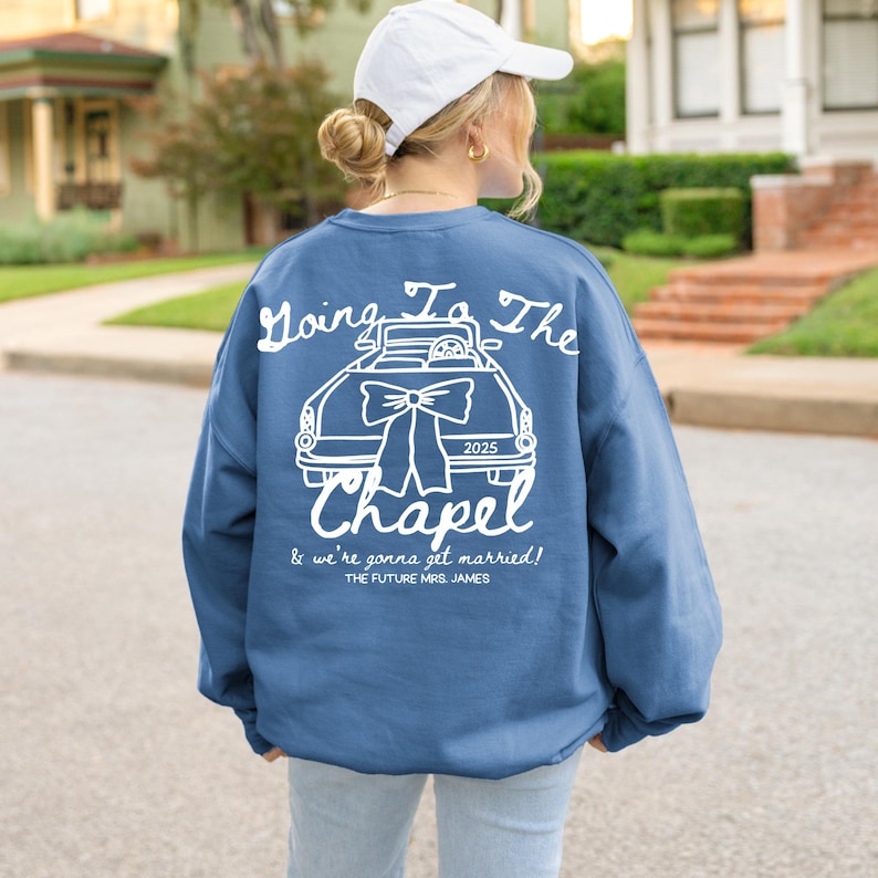 以下が含まれることがあります： 白のテキストと車のグラフィックが特徴の青いスウェットシャツ。「Going To The Chapel」と「2025」、「& we're gonna get married! THE FUTURE MRS. JAMES」と書かれています。カジュアルなスタイルです。