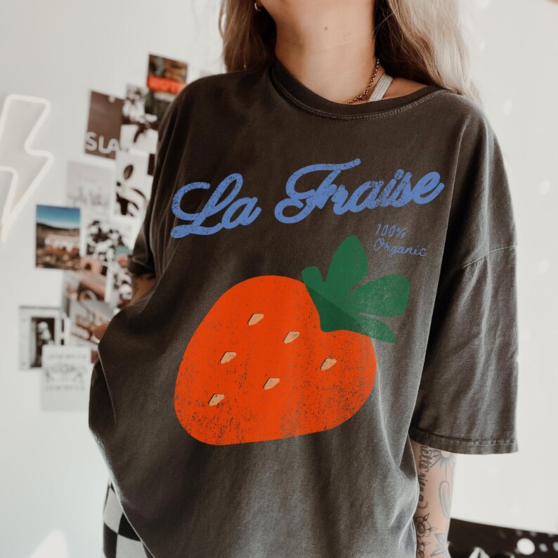 May include: Dark grey t-shirt with a large orange strawberry graphic and green leaves. The text "La Fraise" in blue script is above the strawberry, with "100% Organic" below.