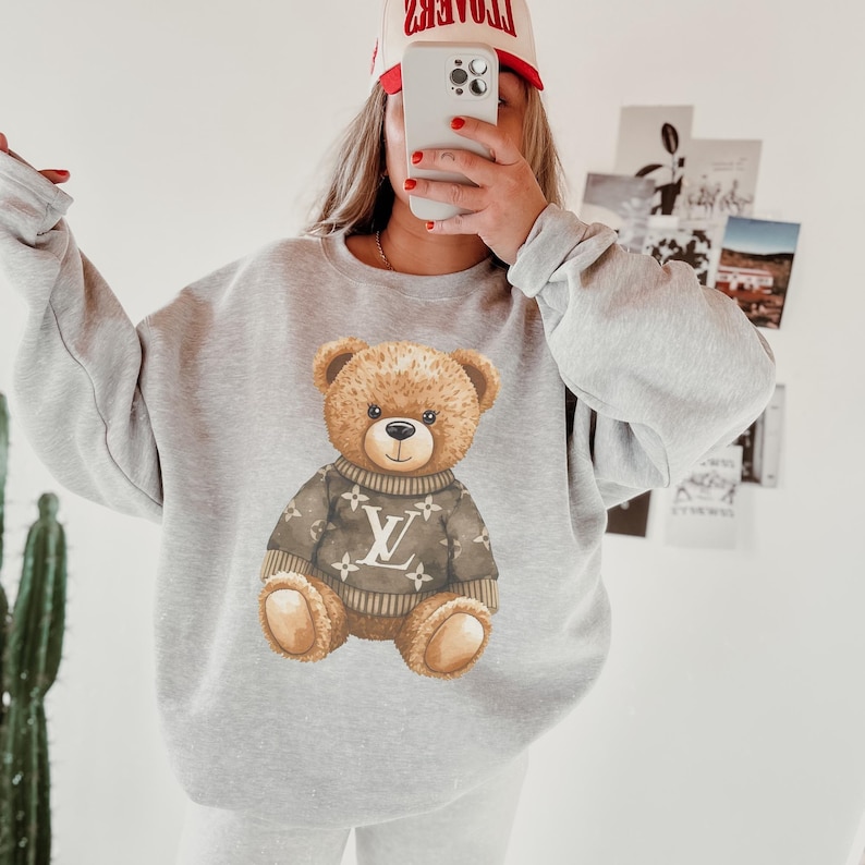 May include: A light gray sweatshirt featuring a watercolor illustration of a teddy bear wearing a brown sweater with a white logo. The bear has brown fur and is centered on the front of the sweatshirt. The person in the image is wearing a red and white hat.