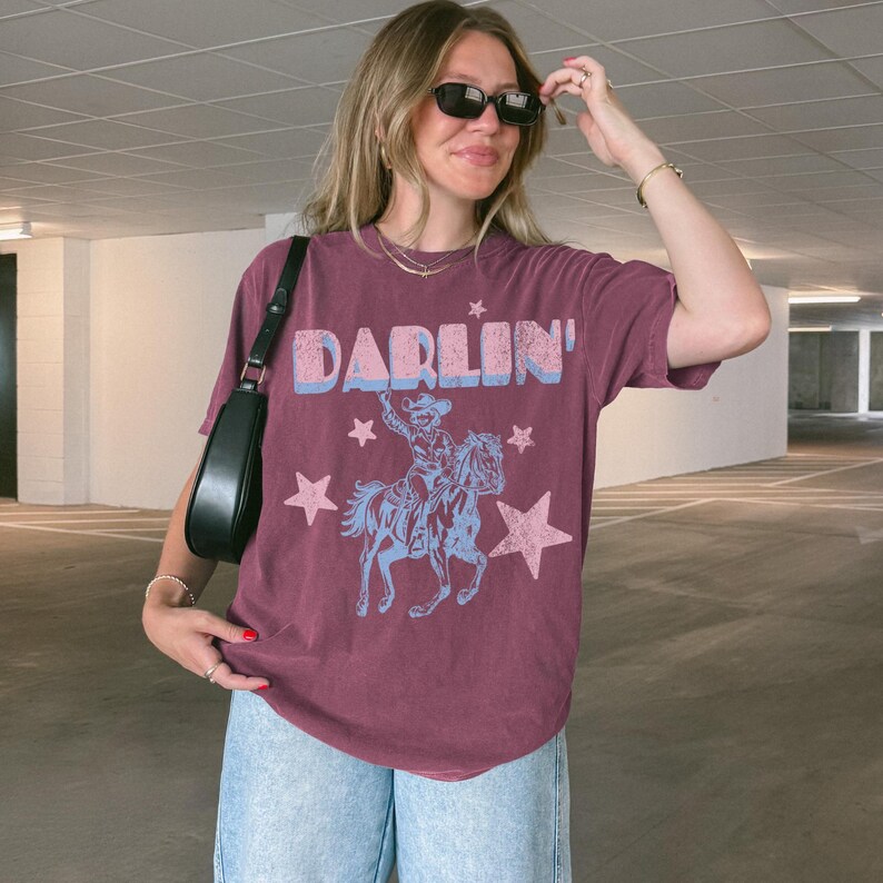 Puede incluir: Camiseta granate con la palabra "DARLIN'" en letras azules y rosas. Debajo, un gr&aacute;fico de un vaquero montando a caballo, tambi&eacute;n en azul y rosa, con estrellas rosas. La camiseta se lleva con vaqueros azul claro.
