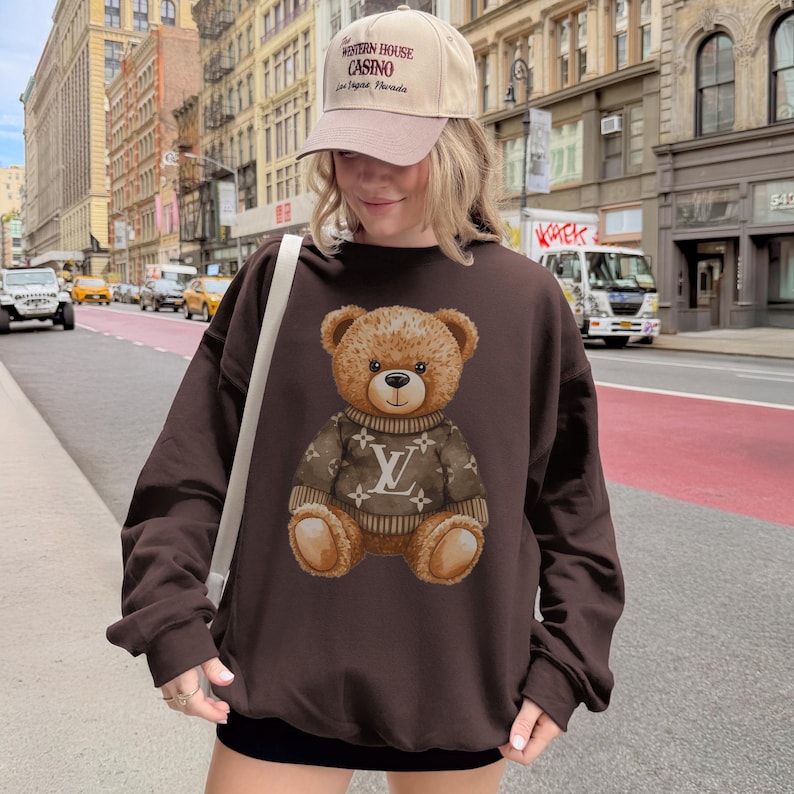 May include: A brown sweatshirt featuring a teddy bear wearing a sweater with a logo. The teddy bear is centered on the sweatshirt. The person in the image is wearing a beige baseball cap with text.
