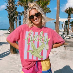 May include: A pink t-shirt with the word "Lucky" in green script and a green duck graphic. The shirt has white and light purple vertical stripes. The person is wearing sunglasses and colorful shorts.