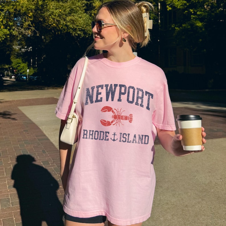 May include: A light pink t-shirt with the text "NEWPORT RHODE ISLAND" in navy blue, and a red lobster graphic. The shirt is worn by a person holding a white purse and a coffee cup.