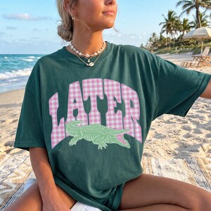 May include: Dark green t-shirt with the word "LATER" in pink gingham lettering and a green alligator graphic. The shirt is short-sleeved and has a relaxed fit. The person is wearing a necklace.