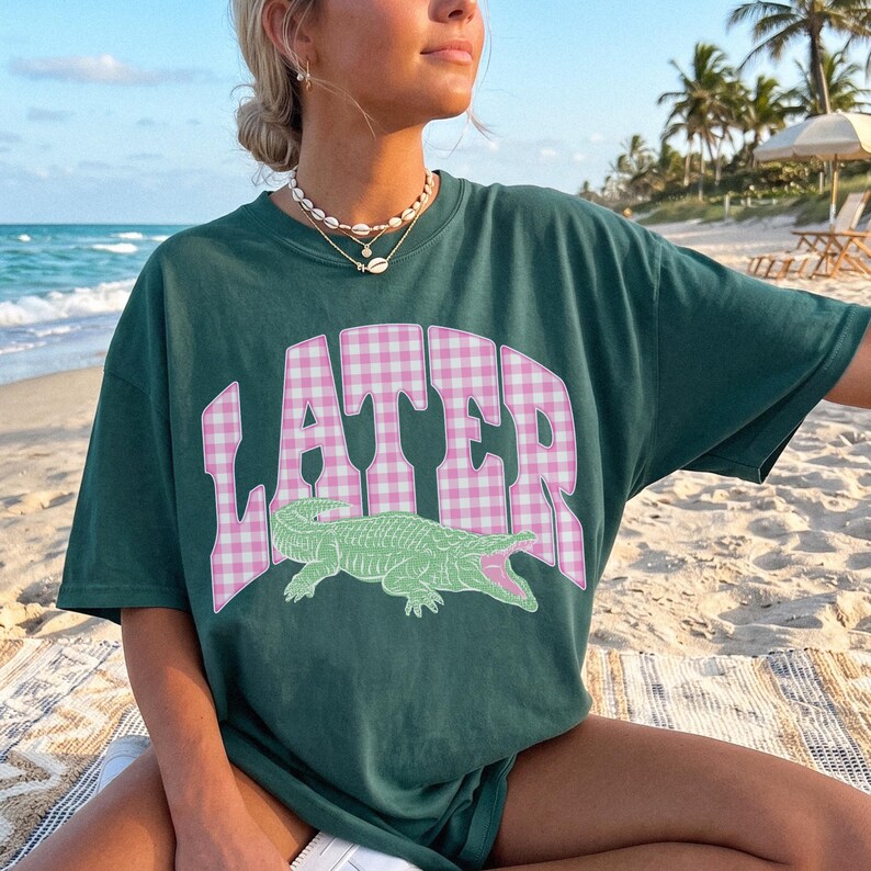 May include: Dark green t-shirt with the word "LATER" in pink gingham lettering and a green alligator graphic. The shirt is short-sleeved and has a relaxed fit. The person is wearing a necklace.