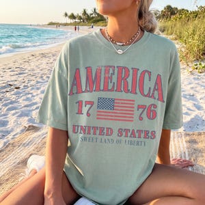 May include: Sage green t-shirt featuring the word "AMERICA" in red, with "1776" and a US flag graphic. The text "UNITED STATES" and "SWEET LAND OF LIBERTY" are printed below. The shirt is worn on a beach.