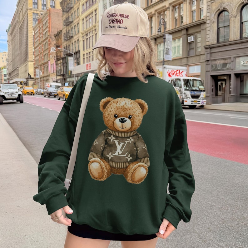 May include: A dark green sweatshirt featuring a teddy bear wearing a brown sweater with a logo design. The teddy bear is centered on the sweatshirt. The person in the image is wearing a beige cap with text.