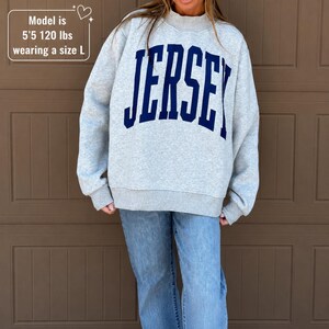 May include: A light gray crewneck sweatshirt featuring the word JERSEY in large, dark blue, arched block lettering across the chest. The garment has long sleeves and a ribbed collar, cuffs, and hem. Text on image: Model is 5'5 120 lbs wearing a size L.