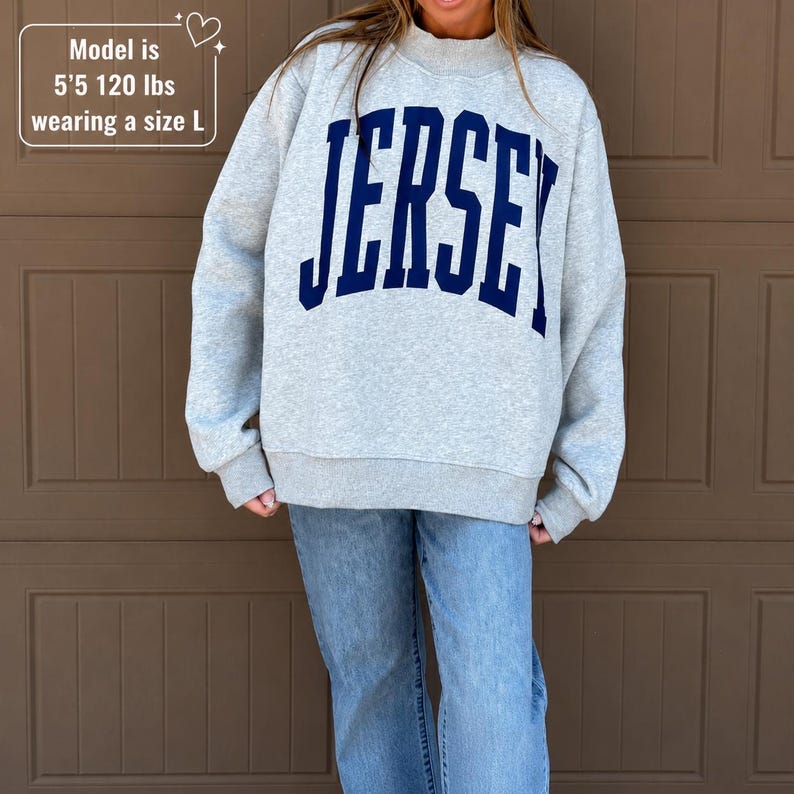 May include: A light gray crewneck sweatshirt featuring the word JERSEY in large, dark blue, arched block lettering across the chest. The garment has long sleeves and a ribbed collar, cuffs, and hem. Text on image: Model is 5'5 120 lbs wearing a size L.