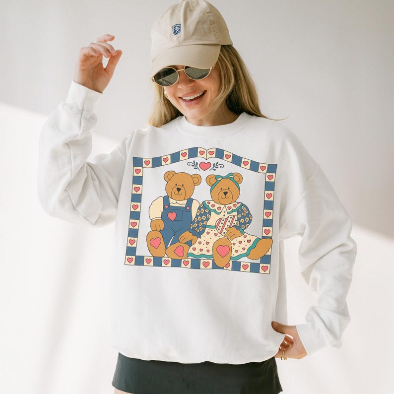 May include: White sweatshirt featuring a framed illustration of two teddy bears. One bear wears blue overalls, the other a floral dress. The frame is decorated with hearts and a blue border. The sweatshirt has long sleeves.
