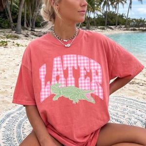 May include: Coral-colored t-shirt with the word "LATER" in a pink gingham pattern and a green alligator graphic. The shirt has short sleeves and a relaxed fit, perfect for casual wear.