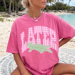 May include: A pink t-shirt with the word "LATER" in a white and pink gingham pattern, and a green alligator graphic. The shirt is a relaxed fit and appears to be made of soft material.