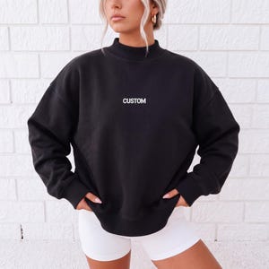 May include: A black long-sleeve sweatshirt with a mock neck collar and ribbed cuffs. The word CUSTOM is printed in white capital letters on the center of the chest. The garment features a relaxed, oversized fit with dropped shoulder seams.
