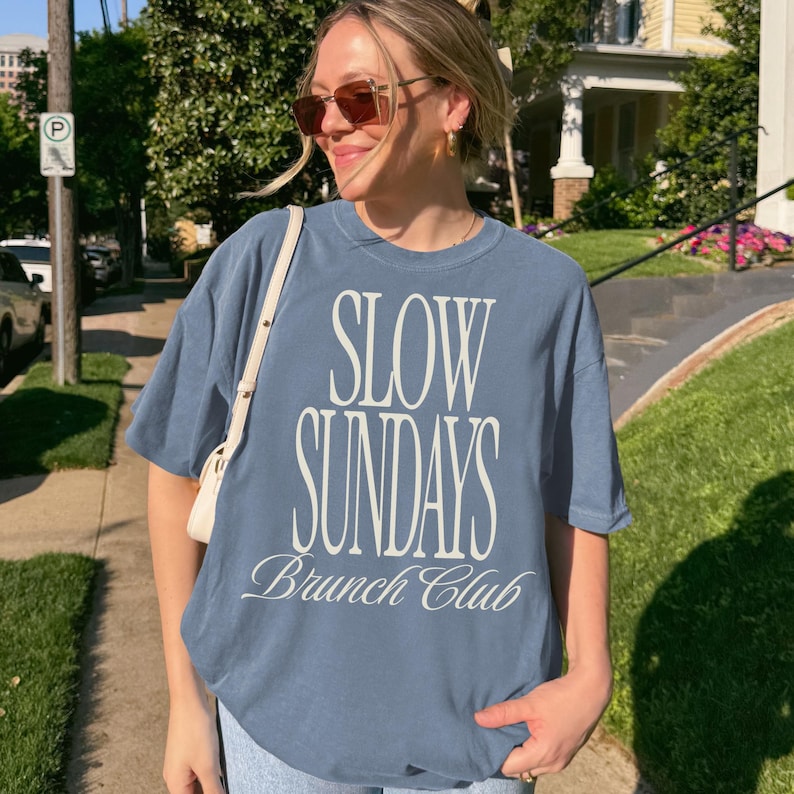 Peut inclure: T-shirt bleu clair avec l'inscription blanche "SLOW SUNDAYS Brunch Club". Le t-shirt &agrave; manches courtes a une coupe d&eacute;contract&eacute;e. La photo est prise &agrave; l'ext&eacute;rieur, par une journ&eacute;e ensoleill&eacute;e.