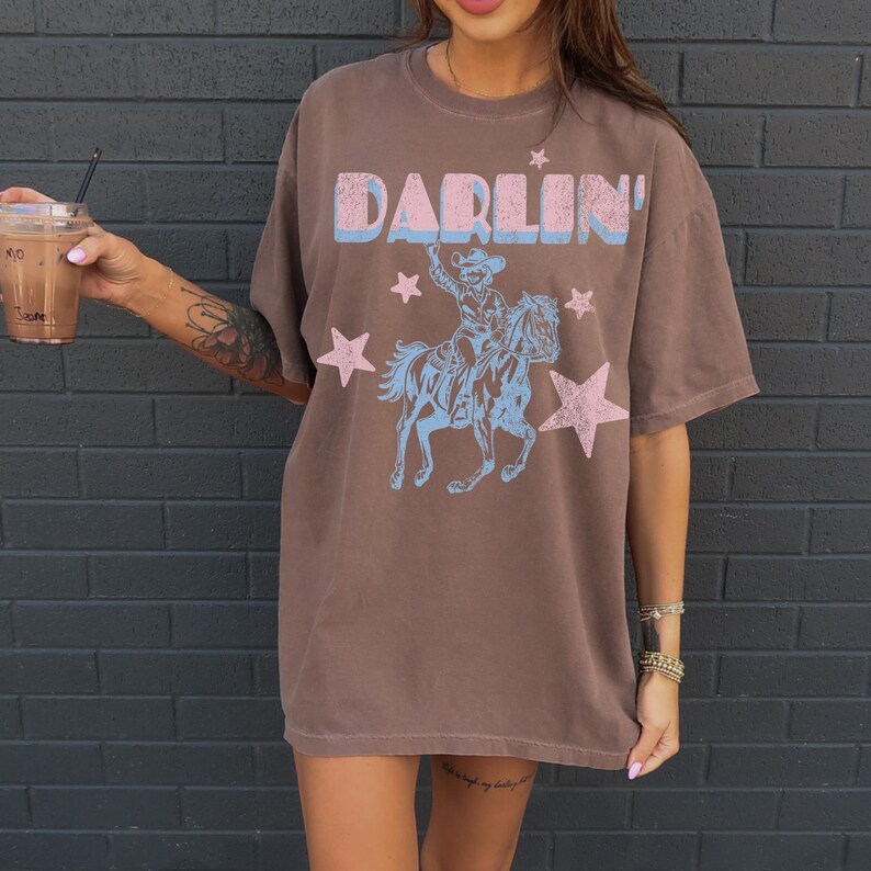 Puede incluir: Camiseta marr&oacute;n con la palabra "DARLIN'" en azul y rosa, un dise&ntilde;o de vaquero a caballo y estrellas. La camiseta tiene mangas cortas y un corte holgado.