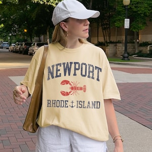May include: A light yellow t-shirt with the word "NEWPORT" in navy blue block letters above a red lobster graphic. Below the lobster, the text "RHODE ISLAND" is printed in navy blue with an anchor symbol. The shirt is worn with a white baseball cap.